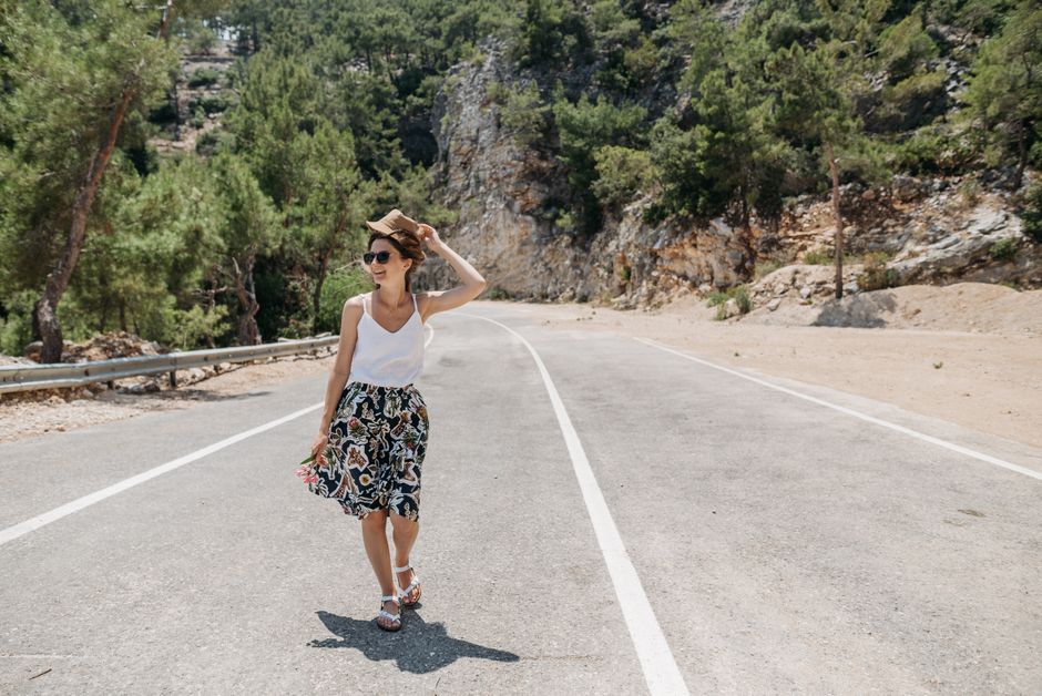 A woman smiling and walking on a sunny road surrounded by trees and nature.