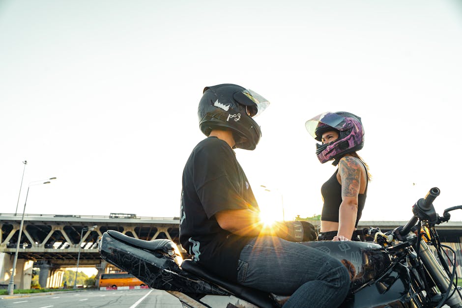 A couple in helmets sit on motorcycles under a city bridge at sunset, capturing a serene urban setting.