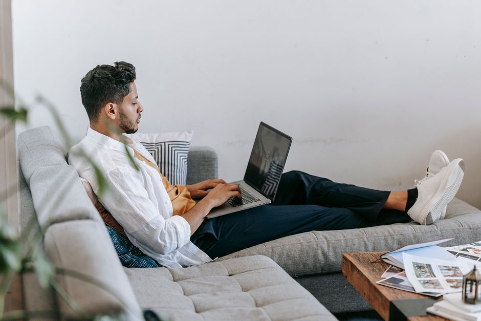 Side view full body of young Hispanic male freelancer sitting on couch and typing on laptop keyboard while working on remote design project at home