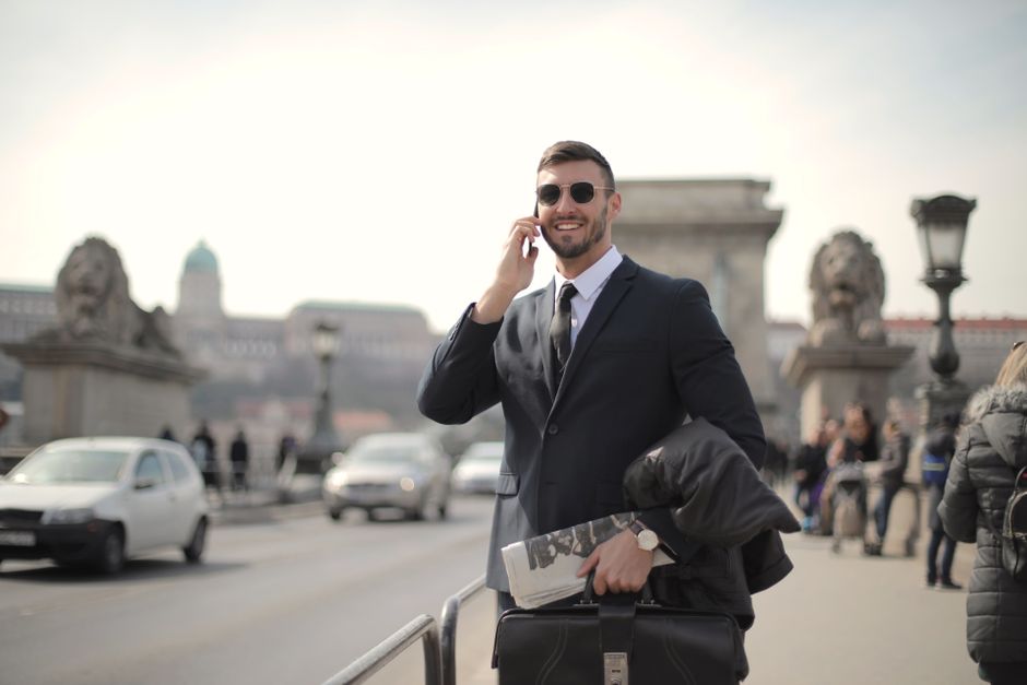 Smiling businessman in formal attire using smartphone on a historic bridge.