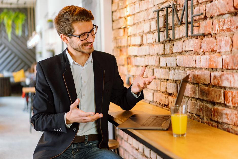 Smiling man in black jacket gesturing during a video call in a modern cafe.