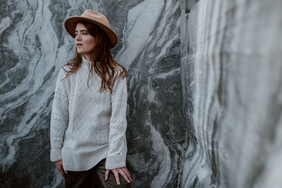 A stylish woman in a fedora hat standing by a marble wall, showcasing fashion in nature.