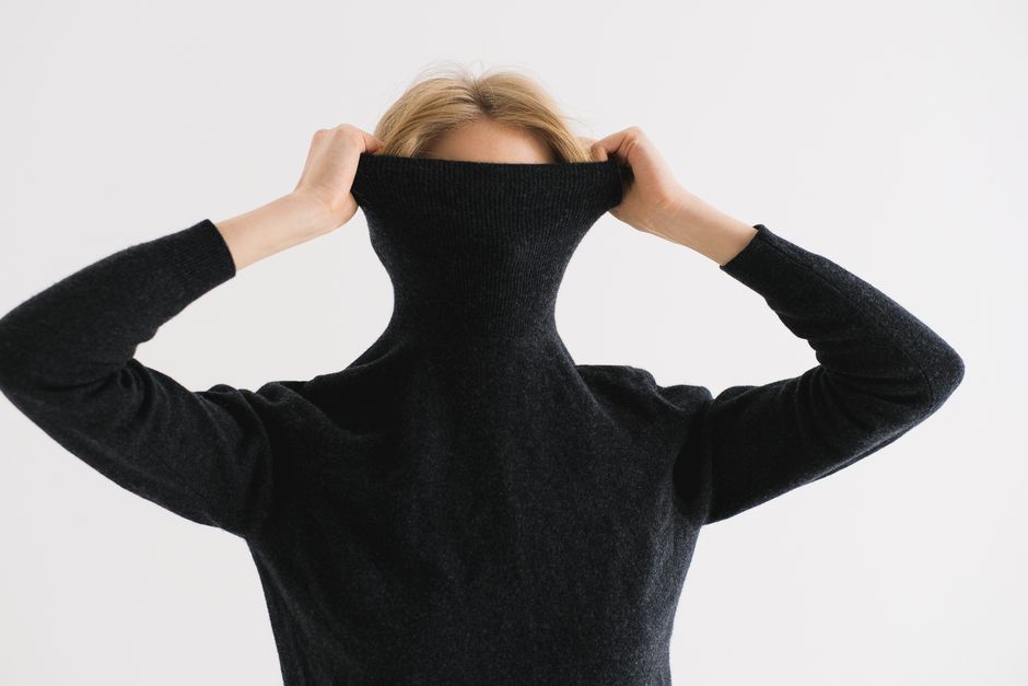 A woman playfully hides her face in a black turtleneck sweater, expressing a shy demeanor.