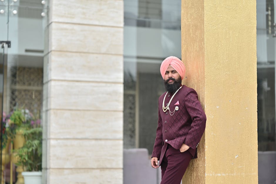 Stylish man in maroon attire with turban and accessories leaning against a gold column outdoors.
