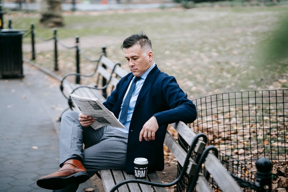 Man in formal attire reading a newspaper on a park bench with a coffee cup.