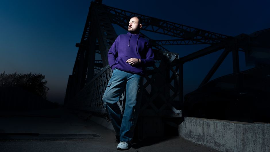 Man in casual attire poses confidently on a steel bridge at dusk.