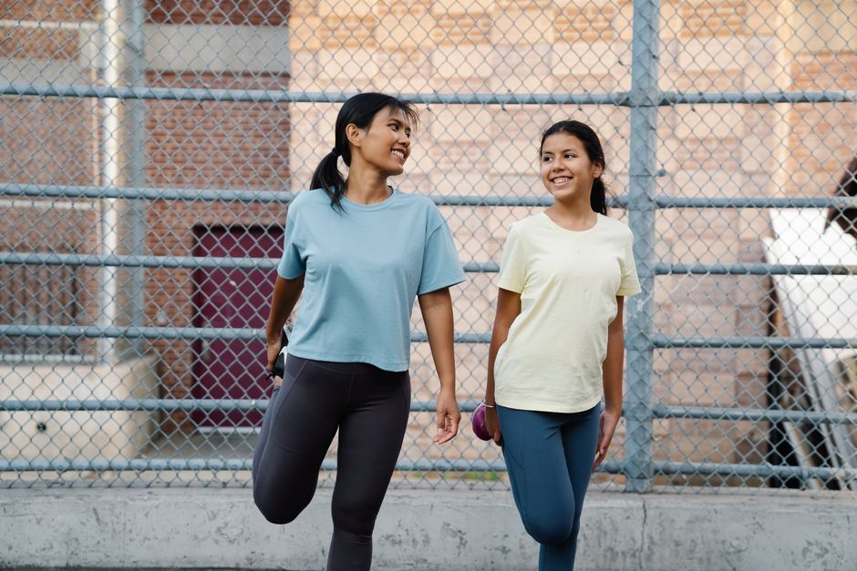 Two women in sportswear stretching outdoors beside a mesh fence, promoting fitness and health.
