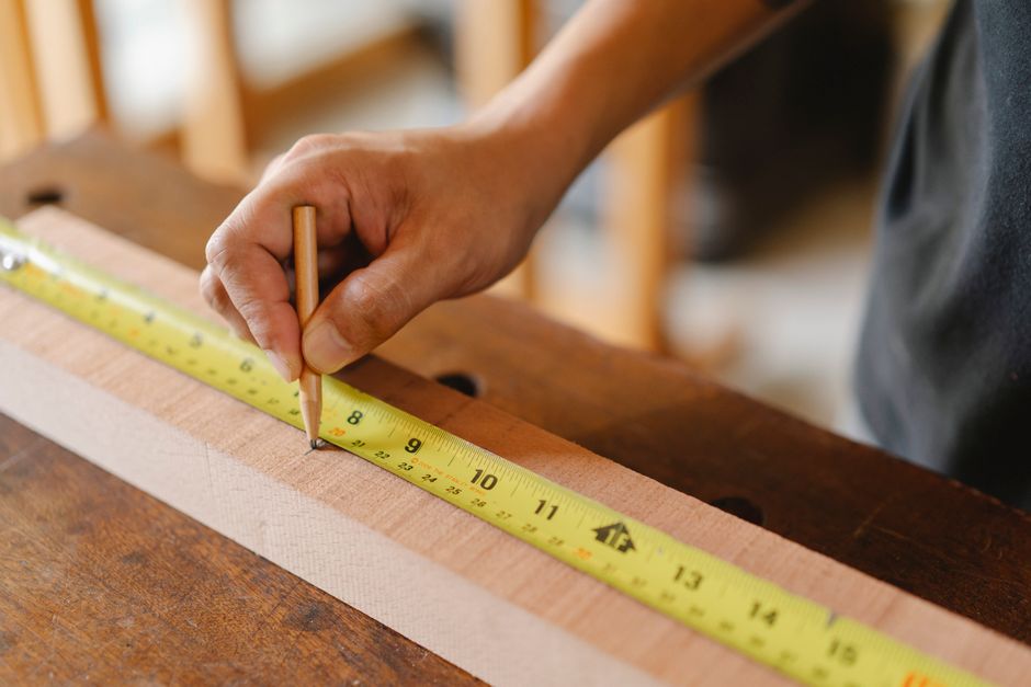 From above of crop anonymous male measuring timber plank and marking size with pencil on blurred background of workshop