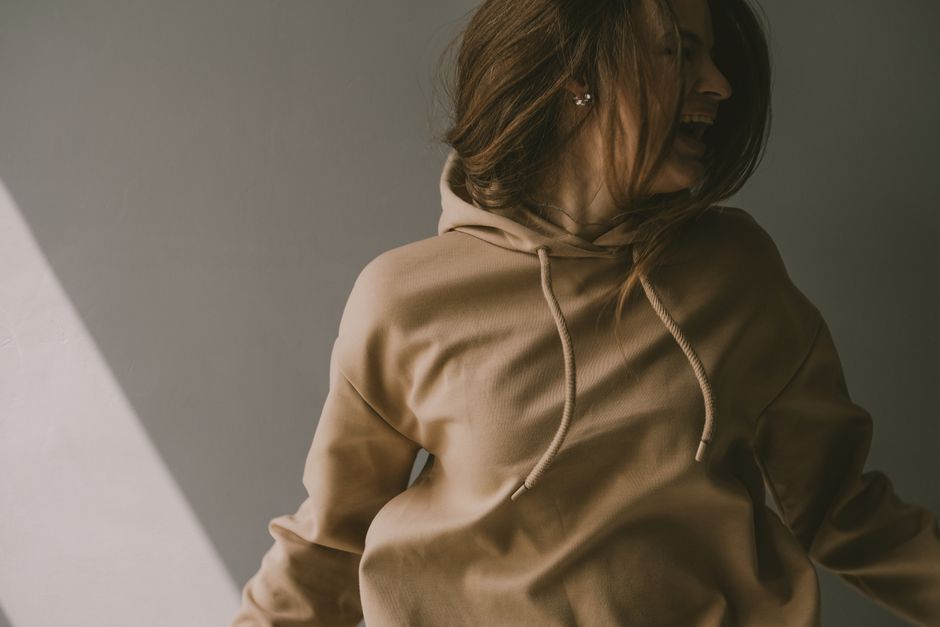A woman joyfully laughing indoors wearing a beige hoodie, capturing a moment of happiness.