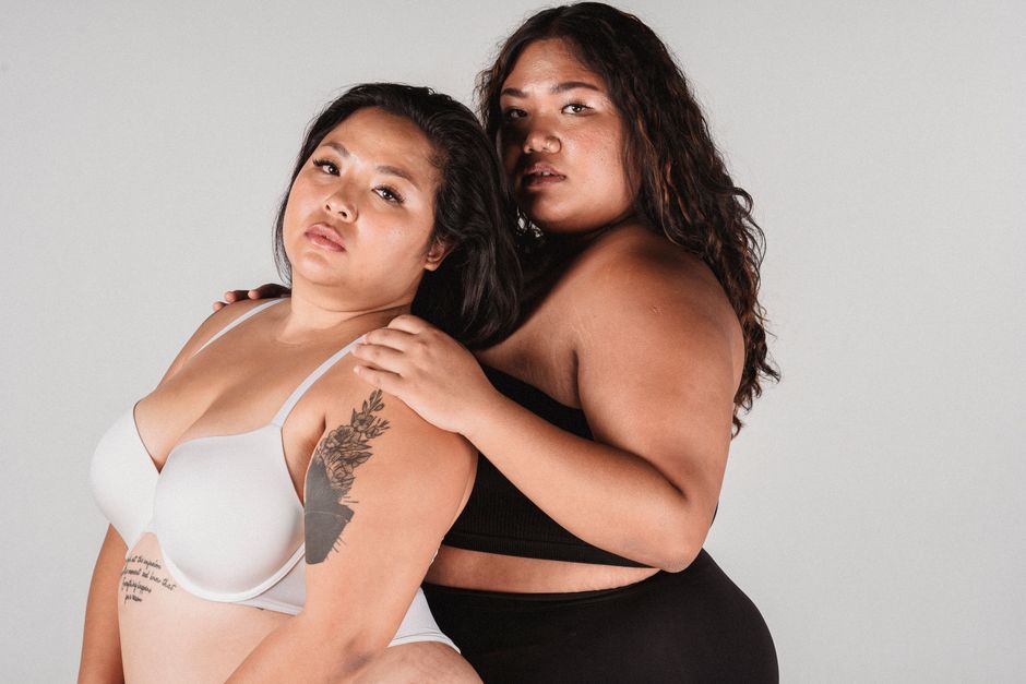 Overweight Asian woman in underwear standing behind back with hands on shoulder of plus size female model while looking at camera