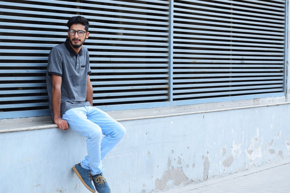 A young man in casual attire sitting by an urban wall with horizontal lines.