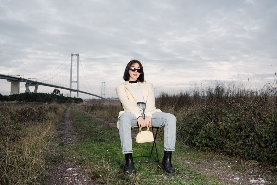 Stylish woman in sunglasses sitting by a bridge, showcasing modern fashion against a natural backdrop.