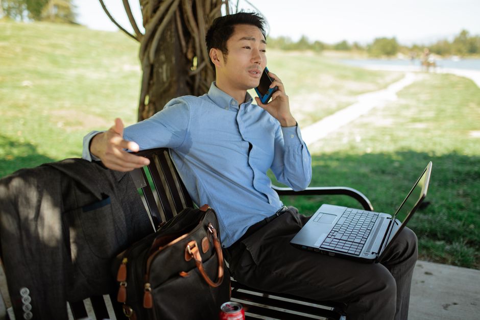 Asian man in business attire working remotely on a laptop, making a phone call in a park.