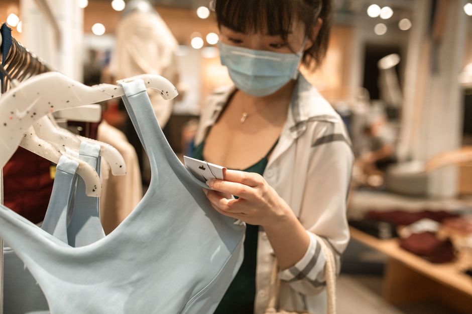 Woman in a store examining clothing tags while wearing a face mask, embracing new normal shopping habits.