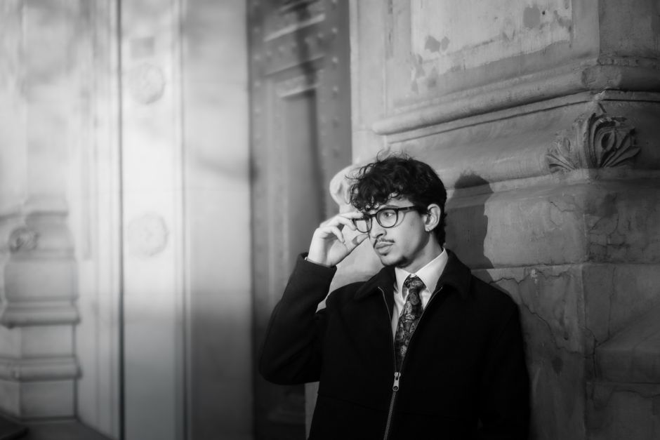 Black and white portrait of a stylish man in an urban setting, wearing glasses and a necktie.