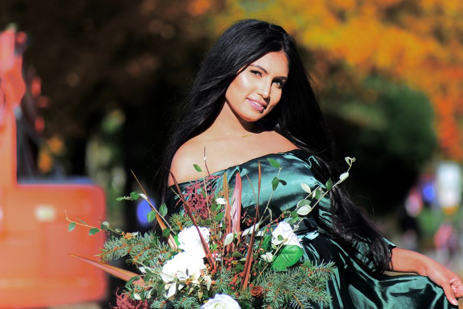 A woman in a green dress poses outdoors with a floral bouquet against a backdrop of fall foliage.