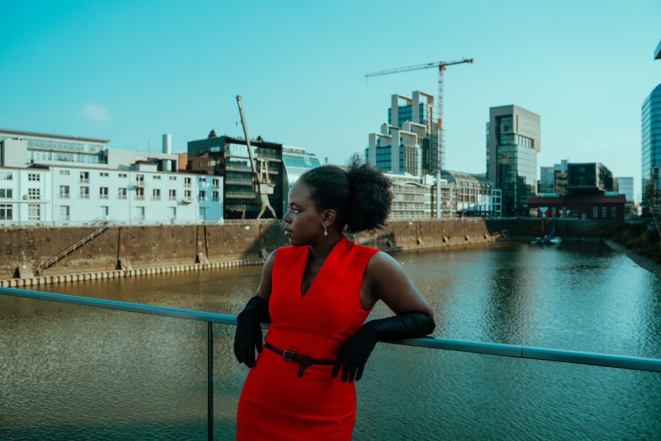 Elegant woman in red dress on a bridge overlooking urban waterfront with modern architecture.