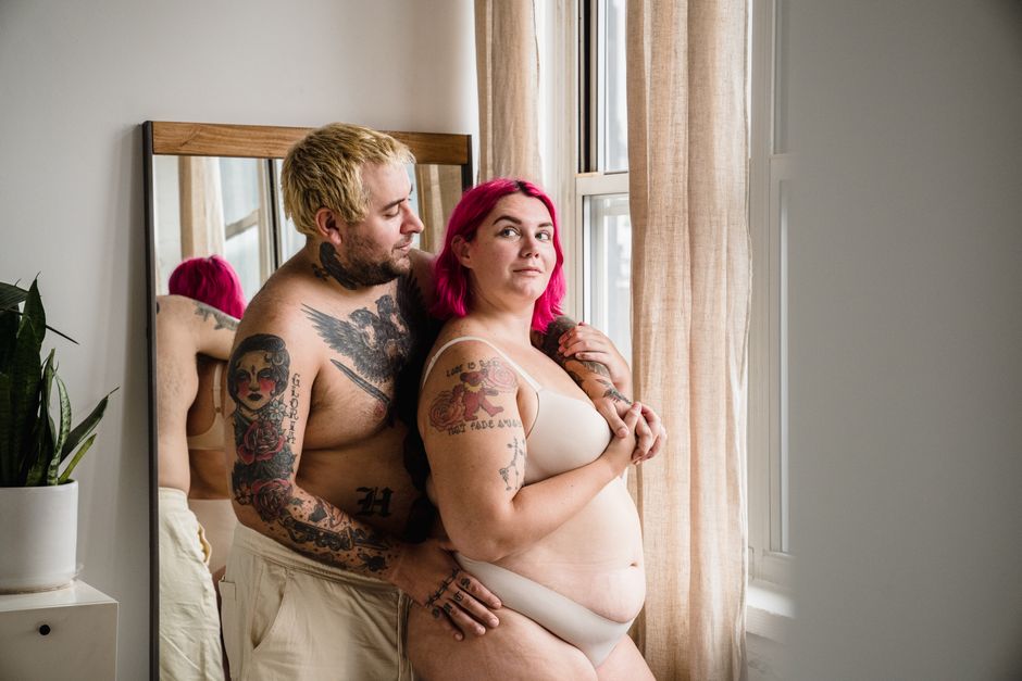 A tattooed couple embracing by a sunny window, promoting body positivity.